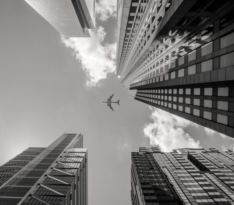 A low angle greyscale shot of an airplane flying above high rise buildings